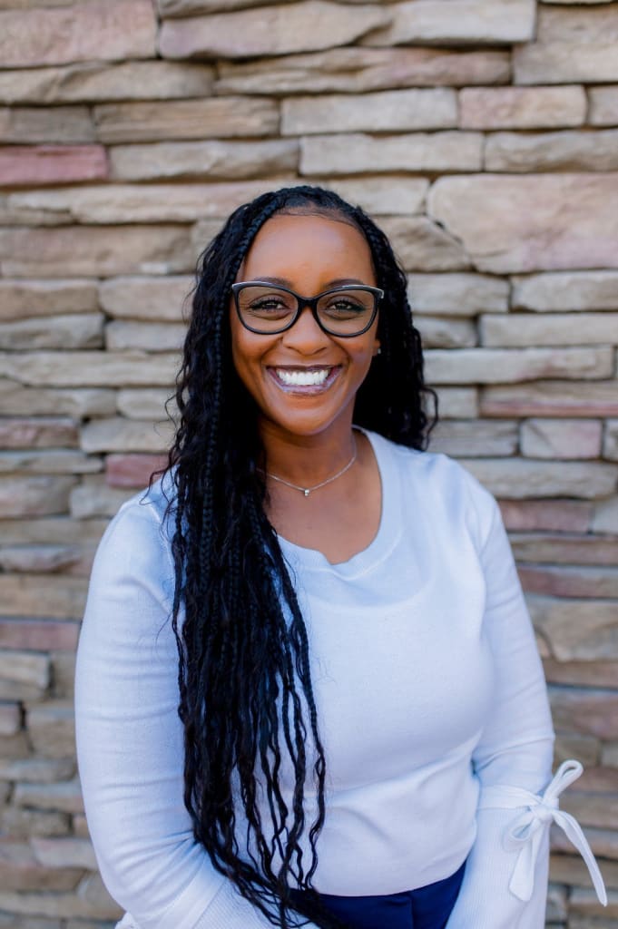 A person with long hair and glasses, wearing a white top, smiles in front of a stone brick wall.